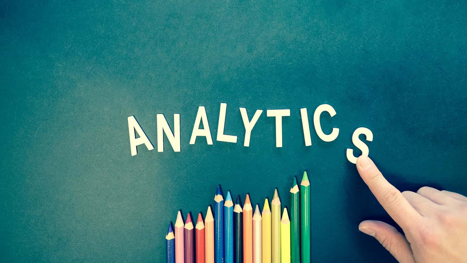 Hand using colored pencils to form the word 'Analytics' alongside digital data icons on a textured background, representing the DIY setup process of Google Analytics for beginners and small businesses